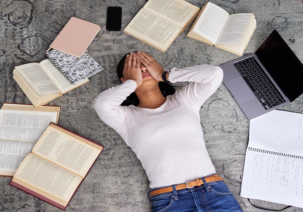 אישה צעירה שוכבת על שטיח ומחזיקה את הראש בייאוש, מסביבה מונחים ספרי לימוד פתוחים ומחשב נייד