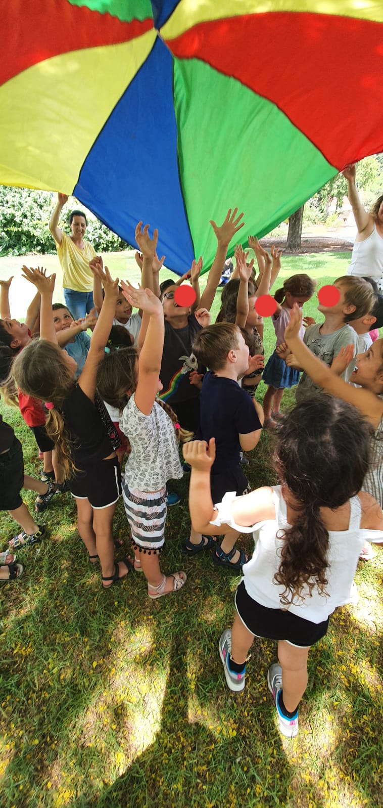 קבוצת ילדים בחוץ עם הדשא משחקים עם מצנח צבעוני גדול מעליהם